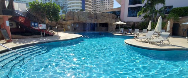 Resort pool deck with pink loungers, white seating pods, ocean view, and palm trees under clear blue sky